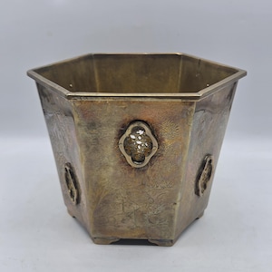 May include: A hexagonal brass planter featuring etched landscape scenes with birds and trees. The vessel has a metallic finish with a warm patina and includes decorative, pierced oval-shaped medallions attached to the exterior panels.