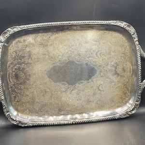 May include: Silver-toned rectangular serving tray with ornate floral and scrollwork detailing. The tray has two curved handles and a decorative rope-like border. The center of the tray has a smooth, oval-shaped area.