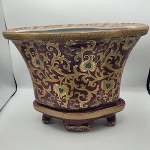 May include: A burgundy ceramic planter with a gold and green floral design. The planter has a flared shape and sits on a matching base with three feet. The rim and base are trimmed with gold.