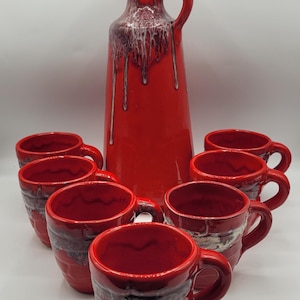 Könnte beinhalten: Ein Set aus roten Keramikbechern und einer Kanne. Die Kanne ist hoch und schlank mit einem Griff. Die Becher und die Kanne haben einen Tropfglasureffekt in Weiß und Dunkelbraun. Ein passendes Set zum Servieren von Getränken.