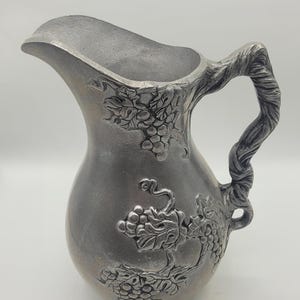 Puede incluir: Una jarra de color peltre con un pico curvo y un asa en forma de vid. El cuerpo de la jarra está decorado con racimos de uvas y hojas. Esta jarra decorativa es adecuada para servir bebidas.