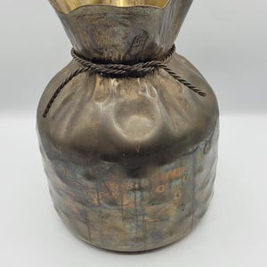 May include: A decorative metal vase with a unique shape, featuring a ruffled top and a drawstring-like detail around the neck. The vase has a metallic finish with a blend of bronze and gold tones, creating a vintage aesthetic.