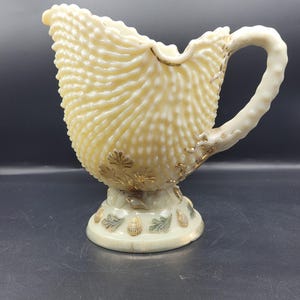 Puede incluir: Cremera de vidrio texturizado color crema, con borde ondulado y asa curva. La cremera presenta detalles de hojas doradas y una base decorativa con diseños de hojas y conchas. Una pieza decorativa de estilo vintage.