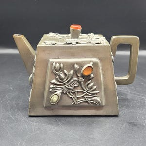 Puede incluir: Una tetera de color plateado con un cuerpo rectangular y un pico. La tetera presenta diseños florales en relieve y está adornada con piedras preciosas naranjas y amarillas. El asa es un simple lazo rectangular. La tapa está rematada con una pequeña gema naranja.