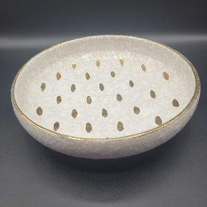 May include: A round ceramic bowl with a textured, off-white glaze and a gold-painted rim. The interior surface features a repeating pattern of small, vertical gold-colored oval shapes arranged in a scattered, uniform grid across the base.