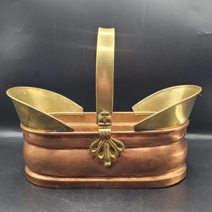Op de afbeelding: Een koperen en messing mand met een handvat. De mand heeft een rechthoekige koperen basis met gebogen zijkanten en een messing handvat. De bovenkant van de mand heeft aan weerszijden twee messing scheppen. Een decoratief messing accent zit aan de voorkant.