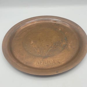 May include: A copper-colored, oval-shaped tray with a raised rim and the word "AFRICA" etched into the surface. The tray has a slightly distressed appearance, with some areas showing a darker patina. The design suggests a vintage or antique aesthetic.