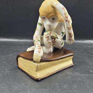Könnte beinhalten: Keramikfigur eines Affen, der auf einem Buch sitzt. Der Affe hat hellbraunes Fell und florale Muster. Das Buch ist braun mit vergilbten Seiten.