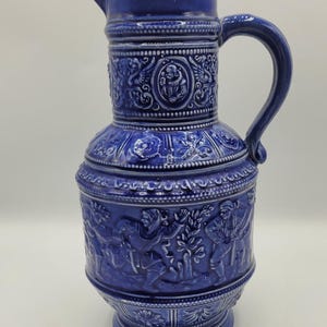 May include: A cobalt blue ceramic pitcher with a handle and spout. The pitcher features detailed relief carvings of figures and decorative patterns around the body and neck. The base is a pedestal design.