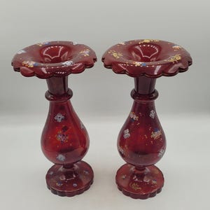 Puede incluir: Dos jarrones de cristal rojo rubí con bordes ondulados y detalles florales. Los jarrones tienen un cuerpo bulboso y una base de pedestal. Los bordes y los cuerpos están decorados con pequeños patrones florales coloridos. Los jarrones miden unos 25 cm de altura.
