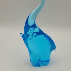 May include: A vibrant, blue glass elephant figurine with a curved trunk and raised ears. The sculpture is a solid, transparent blue with a lighter blue trunk. A decorative piece for home decor.