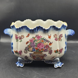 May include: A white ceramic footed bowl with a scalloped rim and two side handles. The exterior features a central floral arrangement in a decorative vase, surrounded by small butterflies and floral sprigs in pink, orange, and blue tones.