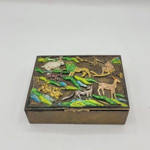 May include: A rectangular, bronze-colored box with a detailed relief scene of various animals, including a lion, deer, and rabbit, set against a backdrop of green and blue foliage. The box is likely used for storage.
