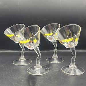 May include: Four clear cocktail glasses with a unique design. Each glass features a slanted bowl, a curved stem, and a round base. The rim of each glass is decorated with a yellow band and a cartoon face. The glasses are arranged on a dark surface.