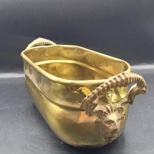 May include: A vintage brass planter with an oval shape and decorative handles. The handles feature a ram's head design. The planter is a warm gold color and appears to be made of solid brass. It is likely used for holding plants or flowers.