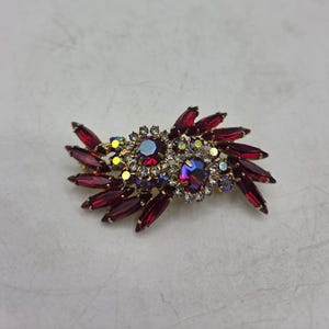Pode incluir: Um broche dourado com cristais vermelho-escuros em lapidação marquise dispostos em leque. O centro é acentuado por duas pedras redondas iridescentes vermelhas maiores, rodeadas por cristais redondos mais pequenos, claros e amarelados.