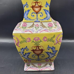 May include: A yellow ceramic vase with a square base and a flared top. The vase is decorated with floral and scroll patterns in pink, blue, and brown. The vase has a height of approximately 10 inches.