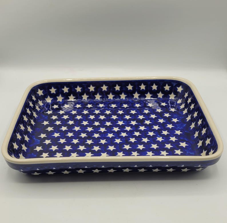 Op de afbeelding: Rechthoekige keramische ovenschaal met een marineblauwe achtergrond en een patroon van witte sterren. De schaal heeft een cr&egrave;mekleurige rand en is geschikt om in te bakken en te serveren. De schaal is ongeveer 30 cm lang en 20 cm breed.