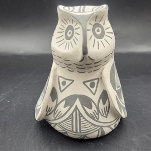May include: A ceramic owl figurine in a sitting position. The owl is primarily white with gray accents. The eyes are large and round, and the wings and body are decorated with geometric patterns. The owl has a stylized appearance.