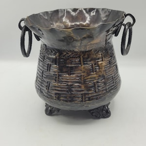 May include: A bronze-colored decorative basket with a woven texture. The basket has a wide, irregular rim and two loop handles. It sits on three ornate feet.