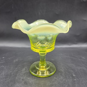 Puede incluir: Un cuenco de cristal con pie que presenta una base de color amarillo vibrante que se transforma en un borde ondulado de color blanco opalescente. El cuenco muestra una superficie texturizada con motivos florales en relieve en el exterior.