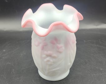 May include: A small, translucent white glass vase with a ruffled, pink-rimmed opening. The body of the vase features a raised, embossed floral pattern in a soft pink hue, showcasing delicate petals and stems against the frosted glass surface.