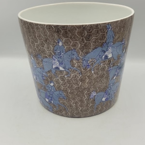 May include: A cylindrical ceramic planter with a brown geometric pattern and blue illustrations of riders on horseback. The interior is white. The planter is suitable for indoor or outdoor use.