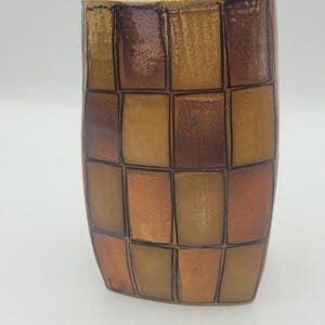 May include: A ceramic vase with a chequered pattern in shades of brown, tan, and copper. The vase has a rectangular shape with rounded edges and a slightly flared opening. The design is reminiscent of a mosaic.