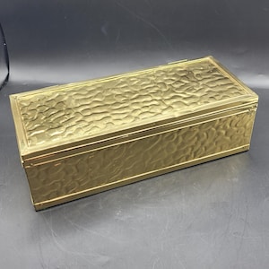 May include: A rectangular, gold-colored box with a textured, hammered surface on the lid and sides. The box has a hinged lid and a simple, elegant design. The box is made of metal and appears to be vintage.