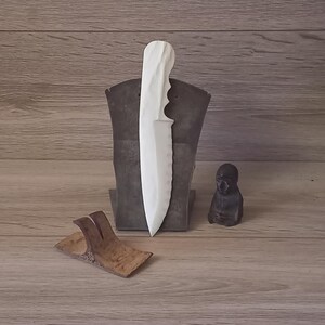 May include: A white knife with a textured blade and handle is displayed on a dark metal stand. A small, dark figurine and a wooden knife rest are also present, set against a wood-grain background.