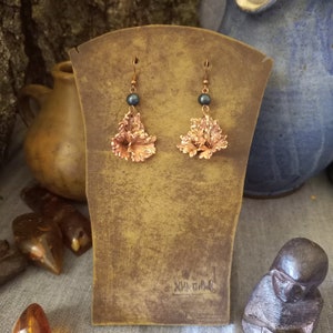 May include: A pair of earrings with a copper flower design and black pearl accents. The earrings are displayed on a brown wooden stand.