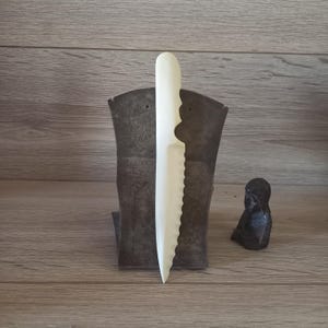 May include: A cream-colored, decorative knife with a serrated edge and a curved handle. The knife is displayed vertically in front of a dark metal stand. A small, dark sculpture is to the right.