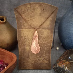 May include: A copper necklace featuring a pendant shaped like a maple seed. The pendant has a textured surface and hangs from a brown chain.