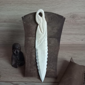 May include: A cream-colored, carved bone letter opener featuring a stylized bird head at the top handle and a serrated, blade-like edge. The handle includes a textured, rope-like detail below the bird head, tapering to a sharp point.