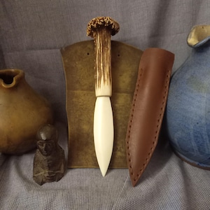 May include: A white-handled knife with a brown antler handle and a leather sheath. The knife is displayed on a brown tile with a small figurine in the foreground.