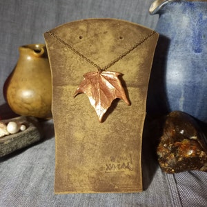 May include: A copper leaf pendant necklace on a brown display stand. The pendant is a detailed, textured leaf shape.