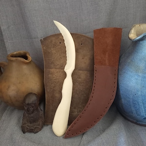 May include: A handcrafted, off-white curved knife with a serrated edge, displayed with a brown leather sheath. The knife is set against a backdrop of pottery and a small statue, suggesting a rustic or artisanal theme.