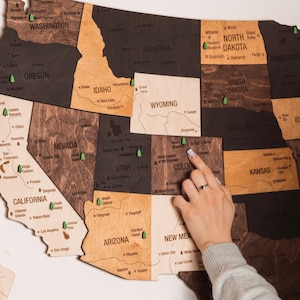 Puede incluir: Un mapa de pared de madera de los Estados Unidos en tonos variados con nombres de estados y ciudades principales grabados con láser, además de pequeños iconos de árboles verdes que marcan parques nacionales. Piezas de madera en tonos marrones.