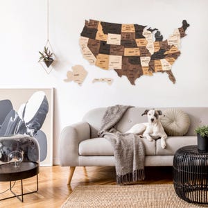 May include: A wooden map of the United States with each state outlined and labeled. The map is mounted on a white wall above a light gray couch with a brown throw blanket. A white dog is sitting on the couch.