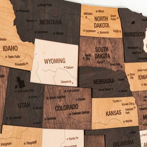 May include: A wooden map of the United States with each state outlined and labeled. The map is made of different shades of brown wood and has a light wood background.