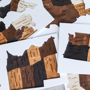 May include: A wooden map of the United States, divided into individual states. Each state is a separate piece of wood, with the state name and some cities labeled. The states are arranged in a grid pattern, with the states in the eastern part of the country on the left side of the grid and the states in the western part of the country on the right side of the grid.