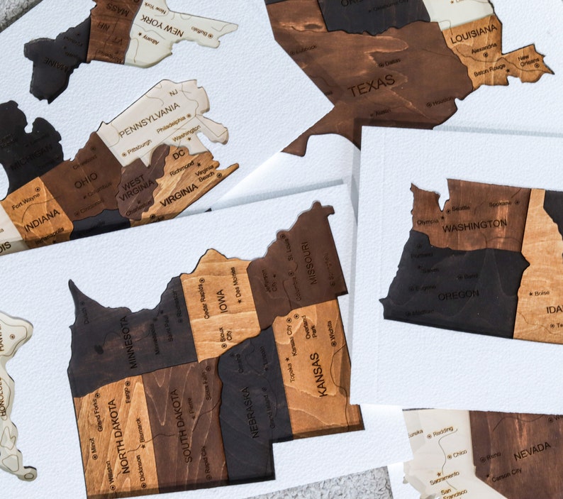 May include: A wooden map of the United States, divided into individual states. Each state is a separate piece of wood, with the state name and some cities labeled. The states are arranged in a grid pattern, with the states in the eastern part of the country on the left side of the grid and the states in the western part of the country on the right side of the grid.