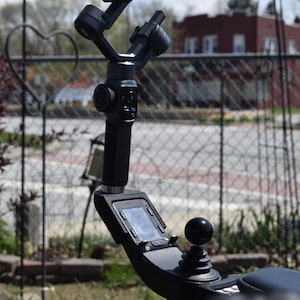 May include: A black camera stabilizer with a small screen and a joystick control. The stabilizer is mounted on a tripod and is pointed towards a blurry background.