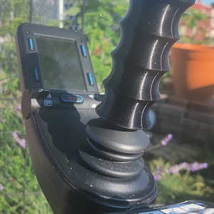 May include: A black joystick with a textured handle and a small screen with buttons. The joystick has a round base with multiple buttons.