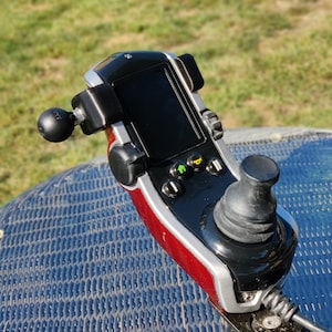 May include: A black and red power wheelchair joystick controller with a black screen, buttons, and a joystick. The controller has a silver trim and is mounted on a metal arm. The background is a blurred green lawn.