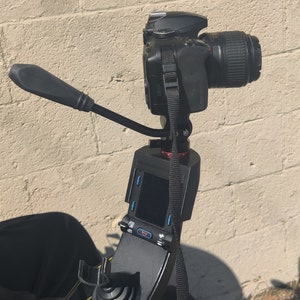 May include: A black camera mounted on a tripod with a remote control panel. The camera is pointed to the right and the tripod is attached to a black chair.
