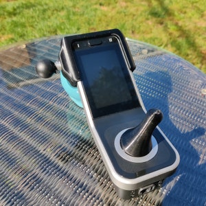 May include: A black and silver handheld device with a screen and a joystick. The device is on a gray and blue surface.