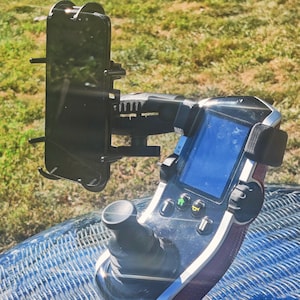 May include: A black phone holder attached to a black and silver control panel with a screen and buttons. The control panel is mounted on a blue surface. The phone holder has a black phone inside.