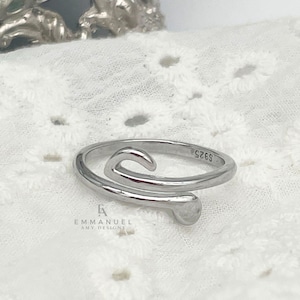 May include: A silver-toned ring with a curved design. The ring has a smooth, polished finish and features a swirling shape. The ring is on a white, textured surface. The ring is marked with "S925".