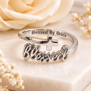 May include: A silver-toned metal ring featuring an open-ended design with a cross symbol and the word Blessed in a cursive script. The interior of the band is engraved with the text Blessed By Grace, showcasing a polished and reflective finish.